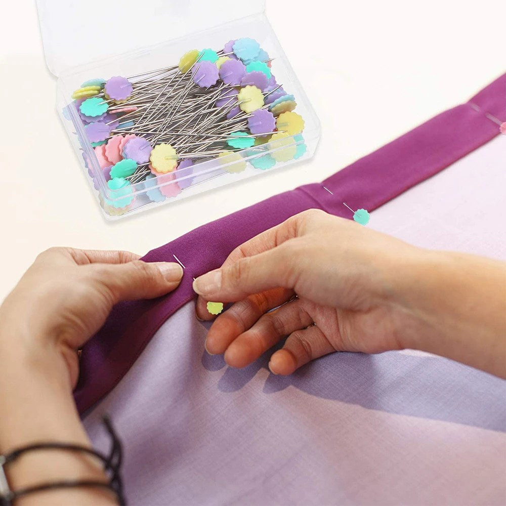 Spilli da sarta con testa a fiore che fermano un tessuto viola, scatola aperta sullo sfondo