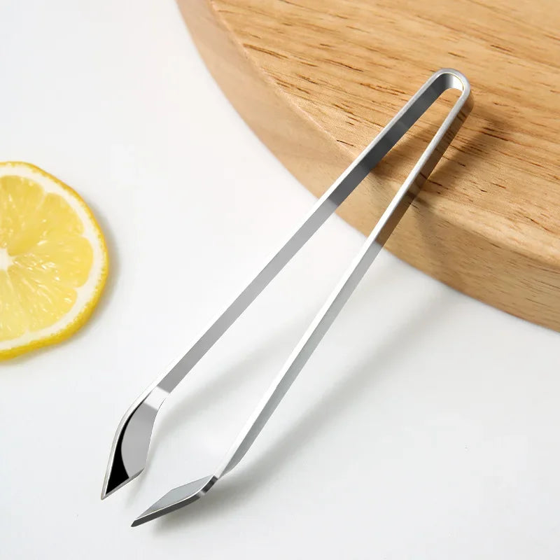 Pinzetta per pesce in acciaio inox con punta obliqua accanto a un tagliere e a una fetta di limone